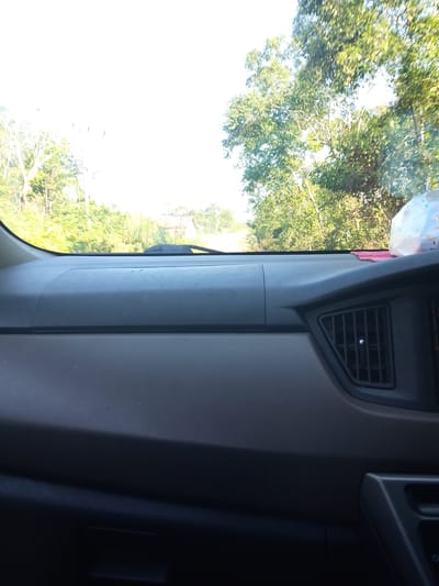 The image shows the interior of a vehicle, looking out through the windshield at a rural road lined with trees. The dashboard is visible in the foreground, with a vent on the right side. The road appears to be unpaved and leads into a heavily wooded area. The sky is bright and hazy, suggesting a sunny day. There are no people or animals visible in the image. It is not possible to determine the exact location in Tiang Tara, Indonesia from this image alone.