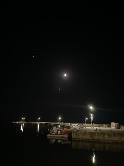 A nighttime photograph captures a waterfront scene in Limenas Avdiron, Greece. The sky is dark with a bright full moon visible towards the upper left quadrant. Several streetlights illuminate a stone-built pier, casting reflections on the calm water below. Small boats are docked along the pier. In the foreground, a white and red inflatable boat is partially visible.
