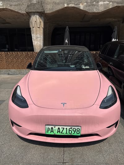 A pink Tesla Model Y is parked on a paved surface in what appears to be an urban setting. The car has a green license plate with the characters "沪A AZ1698". The background shows architectural elements, including a building with a textured façade and arched openings. The reflection in the car's windshield includes black umbrellas. The location is Hongkou District, China based on the "沪A" license plate which corresponds to Shanghai.