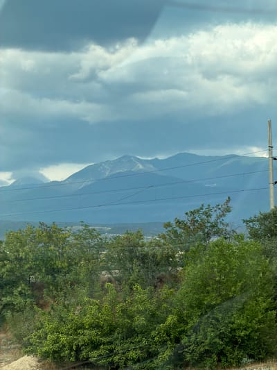 This media file displays a landscape view of mountains, trees, and a cloudy sky. The image is oriented vertically, with the sky and clouds occupying the left side and the mountains and trees on the right. A telephone pole with multiple wires stretches across the midground, partially obscuring the mountain range. The mountains appear distant and are a muted blue-grey color, with some patches of snow or light-colored rock visible on their peaks. The sky is filled with large, fluffy white clouds against a pale blue background, suggesting a daytime scene with possibly changing weather. The foreground is dominated by lush green trees, with dense foliage and some lighter green leaves indicating sunlight. The trees are on the right side of the image, creating a natural frame. Part of a stone wall or embankment is visible at the bottom right corner. There is no visible text or activity occurring in the image. The overall impression is one of a natural, serene landscape, possibly captured from a moving vehicle given the orientation and slight blur. The location context provided is Strumyani, Bulgaria.