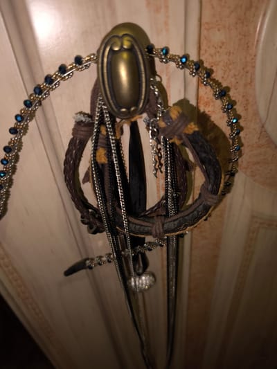 A collection of jewelry is hanging from a hook on a vertical surface. The jewelry includes a beaded necklace with blue and gold beads, several leather bracelets, and multiple silver chains. The beaded necklace is draped over the hook, with the chains and bracelets hanging below. The hook itself is ornate and brass-colored. The background appears to be a light-colored, textured surface, possibly a wall or a cabinet door, with decorative trim. The lighting is dim, creating shadows and highlights on the jewelry. There are no people present in the image. The scene is indoors, in a location that is not discernable as being in the city of Bolshaya Martynovka, Russia.