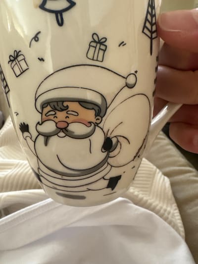 A partial view of a white mug featuring a cartoon depiction of Santa Claus is displayed. The mug is adorned with additional designs, including stylized Christmas trees and gift boxes. An individual's hand is visible, holding the mug by its handle. The background shows light-colored, folded textiles. There is no indication of the mug being in Mikrevo, Bulgaria.