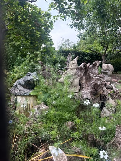 The image shows a close-up view of a garden scene in Leominster, United Kingdom. In the foreground, a stone statue of a frog sits on a tree stump, surrounded by green foliage and small white flowers. Behind the frog, a weathered tree trunk serves as a backdrop for other garden ornaments. These include a stone statue of a gnome, a squirrel, and another small, gnome-like figure. The garden is lush with plants, and the overall impression is one of a whimsical, naturalistic setting. The sky is overcast, suggesting a cloudy day.
