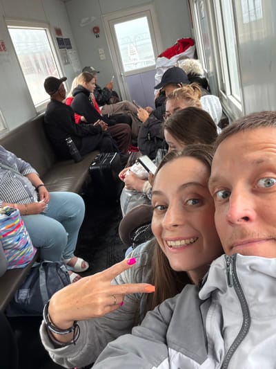 The image captures a selfie taken inside what appears to be a public transport vehicle, such as a ferry or train, in Nizhneleninskoe, Russia. In the foreground, a man with light brown hair and blue eyes smiles faintly, positioned above and slightly behind a woman with long brown hair and blue eyes, who is smiling broadly at the camera. The woman makes a peace sign with her right hand, which features bright pink and orange nail polish. Both are wearing grey and white jackets.

The background shows numerous other passengers seated on rows of dark brown benches. Most are facing forward or looking down, possibly at electronic devices. Several individuals are visible in various casual attire, including jackets and hoodies. The interior of the vehicle has light-colored walls and rectangular windows through which a blurry outdoor scene is visible, suggesting daylight. Luggage and bags are seen on the floor and beside passengers. A red fire alarm and several indistinct signs with unreadable text are mounted on the wall. The overall mood conveyed by the foreground subjects is cheerful and relaxed, indicative of a pleasant journey.