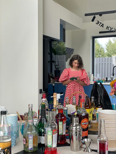 A woman wearing a red and white striped dress stands in a modern kitchen with an island filled with alcohol bottles, paper cups, and a cocktail shaker. She is looking at her phone. A banner with Russian text that translates to "This is the kitchen" hangs from the ceiling. The kitchen has large windows overlooking a backyard with trees and a fence. The scene suggests a party or gathering.