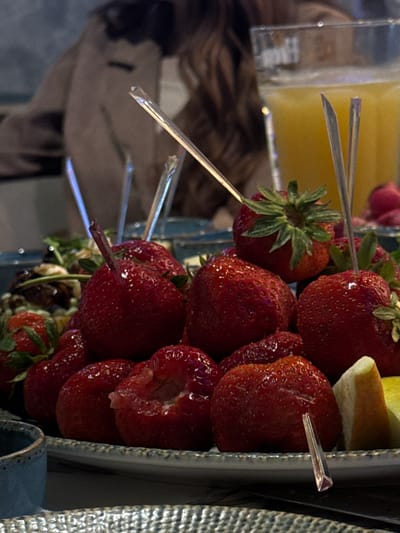 The image depicts a close-up, low-angle view of a serving of fresh red strawberries presented on a plate, presumably within a dining establishment in Zviahel, Ukraine. Numerous strawberries, some whole and plump, others with a bite taken out, are arranged on a white or light-colored ceramic plate with a textured rim. Several clear, triangular skewers are inserted into individual strawberries. To the right of the strawberries, slices of other fruit, including a pale yellow segment resembling a pear and a brighter yellow citrus slice, are visible. In the immediate foreground on the left, a portion of a blue ceramic cup and a textured, possibly metallic or woven, placemat are partially visible.
In the mid-ground, a tall glass filled with a vibrant orange liquid, likely orange juice, stands behind the strawberries. Further in the background, a person with long, dark, wavy hair and a light brown or tan jacket is seated, their upper body partially visible and out of focus, suggesting they are a diner or companion at the table. Other blurry elements in the background indicate additional food items or tableware. The overall lighting is dim and warm, with reflections evident on the glass and skewers, consistent with an indoor evening setting.