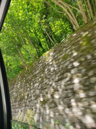 The image depicts a view from inside a vehicle, looking out through a window at a stone wall covered in moss and lichen. The wall runs along a road, and beyond the wall, a dense forest of green trees with dappled sunlight filtering through the leaves is visible. The motion blur suggests the vehicle is in transit. The scene is likely a rural road in or near Golyamo Belovo, Bulgaria, given the context provided. There are no people or specific actions visible, just the natural environment and man-made stone structure.