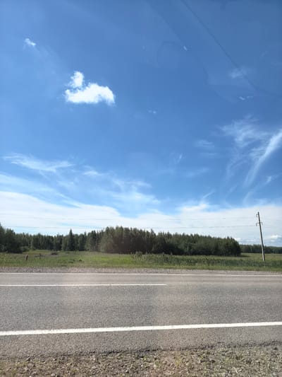 This is a photograph of a roadside landscape under a bright blue sky with scattered white clouds. In the foreground, a paved road with white lane markings runs horizontally across the frame. The road surface appears to be asphalt, with gravel and sparse grass along the shoulder. Beyond the road, there is a strip of green grass and vegetation, followed by a dense line of trees forming a forest. In the distance, a utility pole with wires is visible on the right side of the image. The sky occupies the upper half of the photograph and features wispy cirrus clouds and a few cumulus clouds. The overall impression is of a rural or semi-rural setting, possibly along a highway outside of a populated area. The scene is devoid of any people or vehicles.