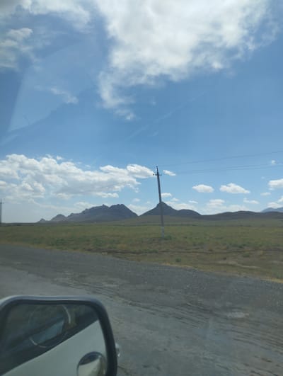 The image depicts a wide, expansive landscape with a clear blue sky and scattered white clouds. In the foreground, a gravel road runs alongside a green grassy field. A power line pole stands tall in the middle ground, with wires stretching across the frame. In the distance, a series of rolling hills and mountains form the horizon. The scene appears to be a rural or undeveloped area, possibly on the outskirts of Asmansay, Uzbekistan, given the open terrain and sparse infrastructure. The weather seems pleasant, with bright sunlight illuminating the landscape. A car's side mirror is visible in the lower left corner of the image, reflecting part of the car's interior and the surrounding environment.