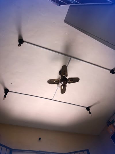The image displays an upward perspective of an indoor ceiling in Bada, Nigeria. The ceiling is off-white, exhibiting minor surface imperfections. A centrally mounted, dark metallic four-blade ceiling fan with an integrated light fixture holding a single white bulb is prominent. Four additional light fixtures, each comprising a dark base and an exposed elongated white bulb, are positioned radially around the central fan, interconnected by exposed silver-colored electrical conduit pipes. Shadows are cast on the ceiling from these fixtures, indicating an overhead light source. A partial view of a light grey, angular structure, possibly a cabinet or air conditioning unit, is visible in the upper right corner, topped by a subtle blue hue. The lower portion of the image shows the top of a light beige wall with a small, dark circular object mounted centrally, and the uppermost edge of a dark metal frame, likely from a window or bed, at the bottom. No individuals or direct actions are depicted.