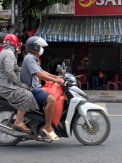 The image depicts a street scene in Tân Quang Commune, Vietnam, with two individuals riding on a white Honda scooter. The person in the front, driving the scooter, wears a grey polo shirt, blue shorts, a dark helmet with a clear visor, and a white face mask. They are carrying two large, orange, sack-like items strapped behind them. The passenger in the back is wearing a long-sleeved grey patterned garment, a red helmet, and a black face covering that extends up to their hairline. The scooter is traveling on an asphalt road, with a white pedestrian crossing line visible.

In the background, a building with a blue and white striped awning covers a storefront. The storefront features dark windows and red tiled walls. A sign with red and yellow lettering is visible above the awning, appearing to be advertising a product. Traffic lights are mounted on a pole to the left of the image, with multiple black signal heads. The environment suggests an urban or semi-urban setting. The overall lighting indicates it is daytime.