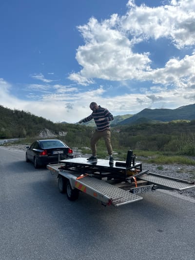 The image shows a man standing on a car trailer parked on a road in Očinići, Montenegro. The man is wearing a striped shirt and khaki pants. He is balancing with his arms outstretched. The trailer has a black car secured on it. The trailer's license plate reads "RP 3368P". The car's license plate reads "KOV 410". The background includes mountains, trees, and a cloudy sky.