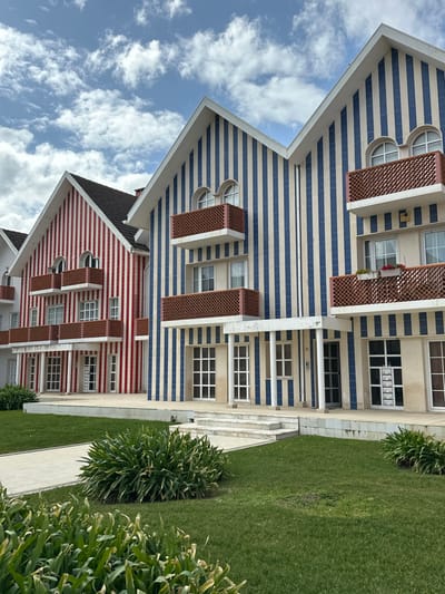 This image displays a daytime exterior view of a row of residential buildings located in Costa Nova, Ílhavo, Portugal. The structures are characterized by distinct vertical striping on their facades; the building on the left is striped red and white, while the two buildings to its right are striped blue and cream. All buildings feature steeply pitched gable roofs. Multiple levels of fenestration are visible, including rectangular windows with white frames and semi-circular arched windows on the upper stories. Balconies with reddish-brown latticework railings project from the facades, supported by white concrete bases. Ground-level entrances consist of white-framed doors and large windows, some approached by white concrete steps and flanked by white cylindrical columns. Minor weathering and discoloration are evident on some white architectural elements. A panel of mailboxes is affixed to the facade of the rightmost building. The immediate environment consists of well-maintained green lawns, several clusters of green shrubs (including broad-leafed plants in the foreground), and paved walkways. The sky above is partly cloudy with scattered white cumulus clouds against a blue background. No individuals or discernable human activity are present in the scene.
