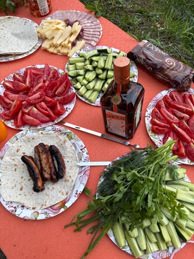 The image depicts an outdoor picnic laid out on an orange tablecloth, likely in the city of Bolshaya Izhora, Russia. Several plates are filled with food, including sliced tomatoes, cucumbers, cheese, and a plate of sausages on tortillas. There is also a bottle of Cointreau liqueur and a package of Delisse chocolate roll. A skewer is visible among the food items. The setting appears to be a casual gathering, possibly a barbecue or a picnic on a grassy area. The overall atmosphere is relaxed and suggests a meal being prepared or enjoyed outdoors.