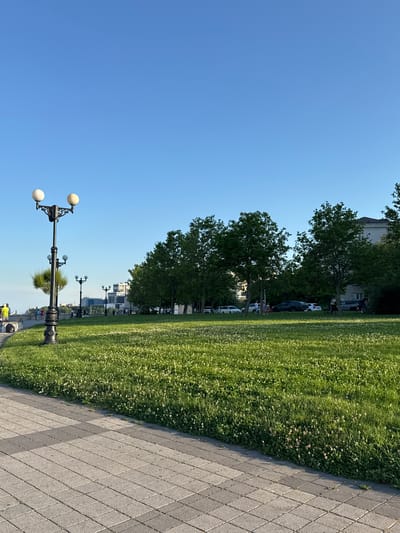 In Neberdzhaevskaya, Russia, a paved walkway runs alongside a lush green lawn dotted with small white flowers under a clear blue sky. A vintage-style lamppost with two spherical lights stands on the left side of the walkway, casting a shadow onto the path. In the background, a line of trees separates the grassy area from a street where several cars are parked. Further beyond the trees, modern buildings rise against the horizon. Several individuals are visible walking along the walkway and near the parked cars, suggesting a public park or waterfront area.
