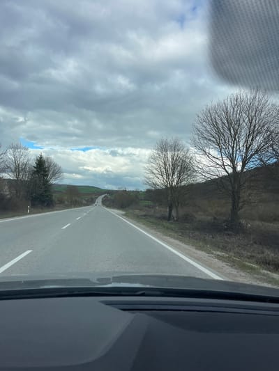A view from inside a vehicle traveling on a narrow road in a rural area. The road has a single white lane marker. On either side of the road, there is uncultivated land with sparse vegetation and bare trees. The sky is cloudy, with patches of blue visible. The road appears to be ascending slightly. The image does not contain sufficient information to confirm the location of Kamburovo, Bulgaria.