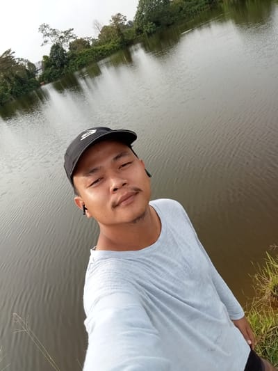 In the city of Kualu, Indonesia, a young man wearing a light blue long-sleeved shirt and a black baseball cap takes a selfie by a body of water. He has dark hair and a mustache. He is also wearing black earbuds. The water, reflecting the overcast sky, takes up most of the frame. In the background, trees and some structures are visible along the shore. The image appears to have been taken outdoors.
