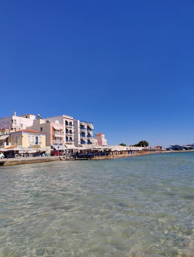 The image depicts a waterfront scene in Aegina, Greece, under a clear blue sky. In the foreground, the shallow, crystal-clear water reveals the sandy bottom. Along the shore, a line of buildings fronts a walkway with numerous outdoor dining areas covered by white awnings. These establishments appear to be restaurants or cafes, with tables and chairs set up for patrons.

The buildings are a mix of architectural styles and colors. On the left, a yellow building with orange trim and blue shutters stands out. Next to it is a taller, white building with a pink section and balconies. Further down the waterfront, a white building with multiple floors and blue-trimmed balconies dominates the view. Several people are seated at the tables, suggesting the area is active.

Further along the waterfront, more awnings extend, indicating a continuous row of businesses. In the distance, a large white yacht is docked, and several dark-colored cars are parked along the waterfront promenade. The overall impression is a vibrant, sunny day in a popular coastal town.