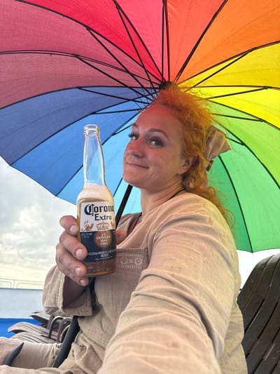 A Caucasian woman with red hair tied back with a beige ribbon is holding a bottle of Corona Extra beer under a rainbow-colored umbrella. She is wearing a light beige, linen-like tunic. She is seated on a dark brown lounger chair. In the background, there are more lounger chairs and a body of water. The sky appears overcast. The image appears to have been taken from a selfie perspective. The location is the city of Belous, Russia.