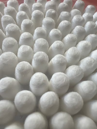 The image shows a close-up, overhead view of a large group of white cotton swabs arranged in a pattern. The swabs have fluffy, rounded tips and appear to be packed tightly together. The lighting is soft and diffused, creating gentle shadows. There is no indication of location in Pskov, Russia. The scene is static and contains only the cotton swabs.