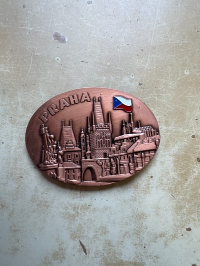 An oval, copper-toned metal souvenir featuring a raised relief cityscape of Prague, Czechia, is depicted resting on a light-colored, scratched surface. The souvenir, oriented horizontally, measures approximately 10-12 cm in its longest dimension. The relief showcases prominent Prague landmarks, including intricate Gothic architectural elements resembling the Old Town Bridge Tower and spires similar to those of St. Vitus Cathedral. Along the top-left curve of the souvenir, the word "PRAHA" is embossed in capital letters. In the upper-right quadrant, a small, enameled representation of the Czech Republic flag (blue triangle on the left, white horizontal stripe on top, red horizontal stripe on bottom) is affixed to the relief. The background surface, possibly a tabletop or counter, exhibits numerous fine scratches, scuff marks, and several small, irregular dark discolorations. No people, actions, or interactions are visible. The object is resting on a surface within the city of Neratovice, Czechia.