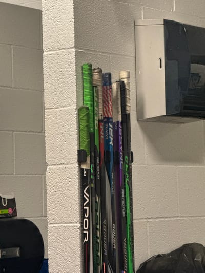 A shelf contains a stack of hockey sticks. Prominent brands include CCM, Bauer, and Vapor. Markings like "P88 / 50 Flex" and "PS2/60 Flex" appear on several sticks, possibly indicating stiffness or blade curve specifications. One stick has an American flag design. Some sticks feature green tape near the top. A white and gray object is visible above the shelf.
