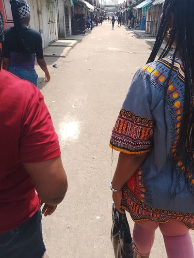 The image captures a vibrant street scene in Mgbuoshimini, Nigeria, characterized by bustling activity under bright daylight. In the immediate foreground, the backs of two individuals are visible, walking away from the viewer. On the left, a person wears a plain red t-shirt and what appears to be blue denim shorts or jeans. On the right, another individual wears a striking grey dashiki top adorned with intricate traditional African patterns in shades of orange, yellow, and red on the sleeves and upper back. This person also has long, dark braided hair, wears a silver-colored bracelet on their left wrist, and carries a dark-colored bag in their left hand.

The paved street stretches into the distance, lined on both sides by numerous simple market stalls or shops, many with closed fronts and corrugated metal roofs. Several other people are visible further down the street, some walking towards the viewer and others away, appearing as silhouettes against the bright backdrop. The overall impression is one of a busy commercial or residential thoroughfare. A distinct, bright reflective patch or glare from the sunlight is prominent on the ground in the mid-distance. The clear blue sky and strong illumination indicate a sunny day. No discernible text is visible in the frame.