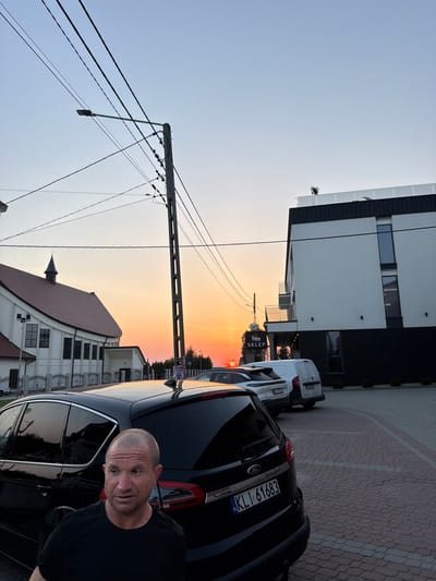 In the city of Mordarka, Poland, a man with a shaved head and a black t-shirt stands in front of a black Ford S-Max. The car's license plate reads "KLl 61683." Behind the car, a modern white and black building with balconies is visible. To the left of the building, a white church with a red roof and a steeple is present. The sky displays a warm sunset with hues of orange and yellow, indicating it is either dawn or dusk. Overhead, utility poles and wires are strung across the sky. Several other vehicles, including a white van and a white SUV, are parked in the vicinity. A sign reading "SKLEP" (meaning "shop" in Polish) is attached to a pole. The ground is paved with reddish-brown interlocking bricks.