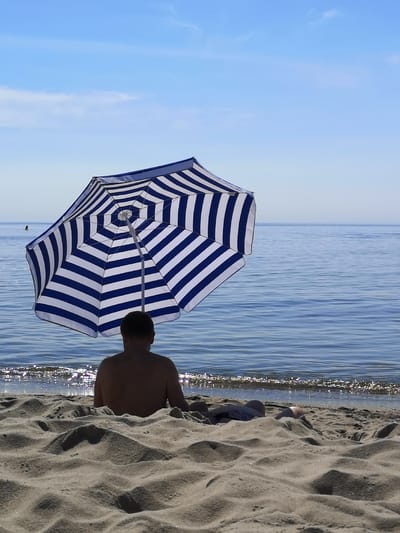 A man sits on a sandy beach, facing the ocean, under a large blue and white striped umbrella. The ocean is calm with gentle waves. The sky is clear blue with a few wispy clouds. The scene appears to be a peaceful day at the beach. The location is the coast of Yantarny, Russia, likely on the Baltic Sea.