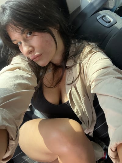 A young woman with dark hair is taking a selfie inside a car. She is wearing a tan unbuttoned shirt over a black tank top, and black shorts. Her legs and thighs are visible in the foreground, and she is leaning back against a dark gray car seat. The sunlight is shining into the car from the right side of the image, creating shadows. The car interior appears to be the setting, and the specific location within Galil Tachton Regional Council, Israel, cannot be determined from the visual information provided in the image.