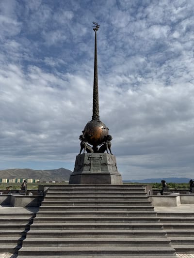The image depicts the Center of Asia monument in Kyzyl, Russia. The monument consists of a bronze globe held by three supporting figures atop a stone pedestal. A tall obelisk rises from the globe, terminating in a stylized crown. The background features a cloudy sky, the Yenisei River valley, and distant mountains. A large staircase leads up to the monument's base. Several cannons are also visible on the upper part of the stairway. Modern buildings can be seen nestled in the valley below.