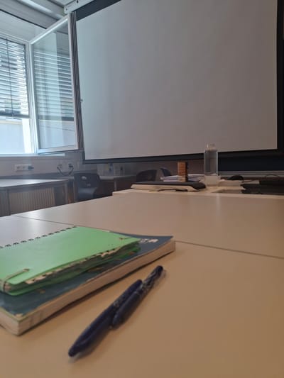 The image depicts a classroom or meeting room setting, likely in Graz, Austria, as indicated by the presence of educational materials and equipment typically found in such institutions. The room features a large projection screen dominating the upper portion of the frame, which is currently blank. A window with venetian blinds is visible to the left, allowing natural light to enter.
In the foreground, a light-colored table surface is set with several objects. Two pens, one predominantly blue with silver accents and another appearing dark blue, lie on top of two notebooks. The top notebook has a green cover, while the one beneath it has a blue cover. Both notebooks appear used, with visible wear and tear, suggesting ongoing activity or study.
Behind the table, in the mid-ground, another table is visible, with a black chair pulled up to it. Electrical outlets and cables are present on the wall behind this table. Further back, other tables and chairs are arranged, and a water bottle stands on a surface near the projection screen. The overall impression is one of an empty, prepared space awaiting a class or presentation.