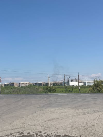 The image shows a bright, clear sky above a landscape featuring power lines, industrial buildings, and lush green trees and grass. A paved road or surface dominates the right side of the frame, showing signs of wear and tear, including tire marks. The scene appears to be set in an urban or semi-urban area, likely in Surenavan, Armenia, given the context.

The power lines crisscross the sky, supported by tall poles, indicating an electrical infrastructure. In the background, several multi-story buildings, possibly apartments or offices, are visible, some appearing to be under construction or in a state of disrepair. Industrial structures, including large silos or storage tanks, are also present, suggesting a manufacturing or processing facility. A plume of smoke or steam rises from one of the industrial areas, indicating ongoing activity.

The vegetation is vibrant and green, suggesting a warm season. The lighting indicates it is daytime, likely late morning or early afternoon, with the sun high in the sky. The overall mood is neutral, presenting a straightforward depiction of the environment. No people are visible in the image. There is a road sign visible at the bottom, but the text is too small to read.