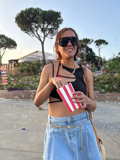 A woman is standing outdoors, holding a red and white striped box of popcorn. She is wearing sunglasses, a black top, and a denim skirt. A gold chain belt with star accents adorns her waist. In the background, trees, a building with lights, and numerous bubbles are visible. The scene appears to be at a park or outdoor event. The location is Sant Vicenç de Montalt, Spain.