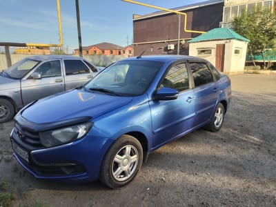 In Kandyagash, Kazakhstan, on a sunny day, a blue sedan is parked on a gravel lot. The car is the primary focus, with its front and side clearly visible. A silver sedan is parked behind it, only partially visible. In the background, a brick building with a light-colored upper story stands next to a small, light-colored structure with a teal roof. Residential buildings with reddish-brown roofs are visible further in the distance, with a power line tower also present. The scene appears to be an outdoor area, possibly a parking lot or an industrial yard.