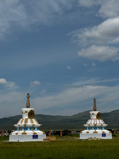 Two ornate stupas stand in a grassy field under a blue sky with scattered clouds. The stupas are white with gold and blue decorative elements. The stupas appear to be identical, with multiple tiered layers and intricate designs. In the background, a line of green hills stretches across the horizon. The ground in the foreground is covered with green grass, and there is a fence with some vehicles behind it. The location is the city of Orongoi, Russia.