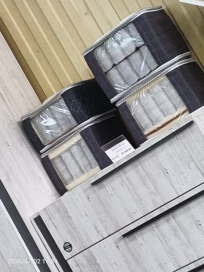 This media file features a retail display of packaged bedding products. The objects are stacked in a way that suggests they are stored on shelves or a similar retail fixture. The background consists of a corrugated metal wall painted a light tan color, and a surface with a wood-grain pattern, possibly a countertop or display stand.
The bedding products are housed in dark fabric bags with clear plastic windows, allowing the contents to be visible. The contents appear to be rolled items, possibly mattresses or large pillows, in a light grey or off-white color. Each bag has a decorative trim along the top edge, consisting of a braided rope pattern in white and black.
One of the packages has a white label attached, displaying text. The visible text reads: "18 100.00". Below this, there's another line of text, partially obscured, that appears to be "ITKHAA".
The image is well-lit, suggesting an indoor setting, likely a store. There are no people visible in the frame. The overall impression is a clean, organized retail environment. The timestamp in the bottom left corner, "2026/07/02 17:34", indicates the date and time the image was captured, suggesting a late afternoon capture.
The location context provided is Chany, Russia. This information, combined with the visual elements, points to a retail store in this specific Russian town. The scene depicts commercial activity, showcasing products for sale.