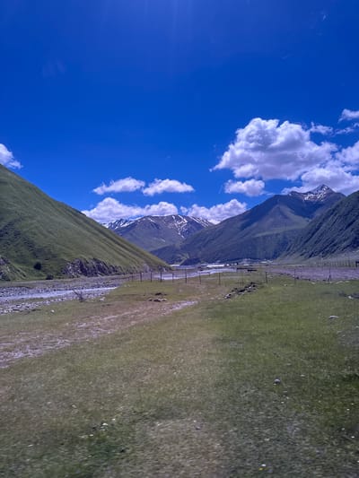 The image shows a wide, panoramic view of a mountainous valley with a riverbed and green, grassy slopes. The sky is a vibrant blue with scattered white, fluffy clouds. In the foreground, there is a grassy meadow with some rocky patches and a dirt path. A shallow, rocky river flows through the middle ground. Further back, the slopes of the mountains are covered in green vegetation, and the peaks are partially covered in snow. There are some fence posts scattered throughout the landscape. In the distance, a small settlement or village can be seen nestled in the valley. The overall scene depicts a natural, rugged landscape, likely in a rural or mountainous region. Without further information or visual cues, it is not possible to definitively place this image in the city of Almasiani, Georgia.