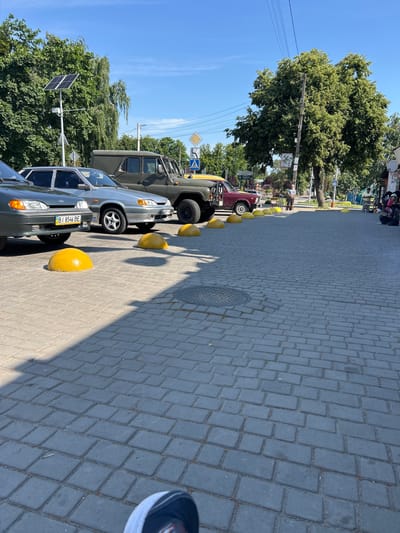 A bright, sunlit scene unfolds on a pedestrian street paved with interlocking gray bricks. The street is lined with parked vehicles, including a dark green military-style SUV, a burgundy car, and two silver sedans. Yellow, dome-shaped concrete bollards are strategically placed in front of the vehicles, likely to prevent parking. In the background, lush green trees with full canopies cast dappled shadows on the ground. A solar panel is mounted on a pole near the trees, indicating a commitment to renewable energy. Traffic signs, including a pedestrian crossing sign and a diamond-shaped warning sign, are visible. A few people are present in the scene, some standing near a storefront and others sitting on a bench, possibly near a bus stop. The overall impression is one of a peaceful, everyday scene in a well-maintained urban environment. The image likely captures a street in the city of Opishnia, Ukraine, a town known for its pottery and traditional crafts.