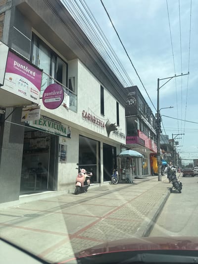 This image depicts a commercial street in Pitalito, Colombia, viewed from inside a vehicle. The scene is dominated by a row of multi-story buildings lining the right side of the street under a partially cloudy blue sky.

The primary building in the mid-ground features a white facade with dark framed windows on the upper floor and large glass storefronts at street level. Two "puntored Corresponsal Bancario" signs are visible: a large rectangular pink sign on the far left edge of the frame, and a smaller circular pink sign mounted on a balcony railing. Below these, a green and white sign identifies "Tex Vicuña Almacén y Sastrería." Inside "Tex Vicuña," a male individual in a light-colored shirt is seated at a table. Adjacent to this, a "Carnicería CARNE MOTTA" (Butcher Shop) is indicated by a prominent sign on the building, featuring a large, dark, three-dimensional bull's head mounted below the name.

On the sidewalk in front of the "Carnicería," a light pink vintage-style scooter is parked. Further down, a smaller blue scooter is parked near an aqua-colored umbrella covering a small vendor's stand. The sidewalk itself is paved with light grey blocks featuring red-painted grid lines. A utility pole laden with numerous overhead electrical wires stands prominently on the right side of the street, extending towards the sky. Further down the street, multiple motorcycles are parked on the sidewalk, and several indistinct figures are visible on the pedestrian walkway. Vehicles are visible further down the street, indicating active traffic. The foreground shows the red hood and windshield of the vehicle from which the photograph was taken.
