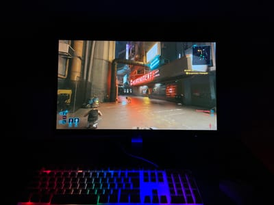 The image displays a computer workstation in a dimly lit indoor setting, presumably located in Enem, Russia. A wide-screen computer monitor is centered, presenting a video game. Below the monitor, a mechanical keyboard with multi-color RGB backlighting is visible, accompanied by a partially obscured dark computer mouse to its right. The surrounding physical environment is dark, revealing no additional details.
The game displayed on the monitor is Cyberpunk 2077, depicting a futuristic, industrial urban environment. A player character, viewed in third-person, is seen crouching in a narrow, neon-lit alleyway. The character is wearing a dark sleeveless top and light trousers. The in-game environment features tall, metallic structures, exposed pipes, and various neon signs, including one partially displaying "ENDIMENT" and another clearly showing "СПАСИБО" (Thank You) in Russian. The ground within the game reflects ambient light from the neon signs. A distant non-player character is visible further down the alley. The game's user interface is present, featuring a minimap in the top-right corner, Russian objective text including "Встретиться с Такемурой" (Meet Takemura) and "Вытащить оружие" (Draw weapon), and player status icons in the lower-left. A red hostile indicator is visible in the lower-right of the game screen.