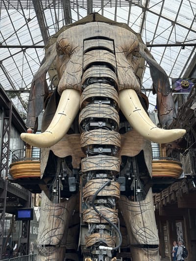 An enormous, wooden, mechanical elephant stands in the Machines de l'île in Nantes, France. The elephant, a colossal automaton, is intricately detailed with carved wood, resembling aged skin and muscle. Its tusks are large, curved, and a creamy white, extending outwards from its head. The elephant's trunk, also made of segmented wood, hangs down, revealing a complex internal structure of metal gears, pistons, and hydraulics. Platforms with wooden railings are visible on either side of the elephant's body, suggesting a viewing area for visitors. The background shows a vast, glass and metal atrium, characteristic of an industrial exhibition space, with glimpses of other mechanical structures and industrial elements. People can be seen at the base of the elephant, dwarfed by its scale, observing the spectacle. The overall environment is one of awe-inspiring, steampunk-inspired engineering.