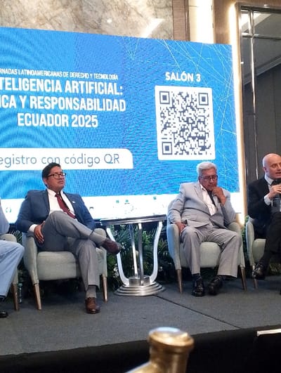 The image displays an indoor conference or panel discussion set on a raised stage within "SALÓN 3" of an event venue in Samborondón, Ecuador. Four adult males are visible, seated in light grey armchairs arranged in a row.

In the background, a large, rectangular blue digital screen features white text that reads: "JORNADAS LATINOAMERICANAS DE DERECHO Y TECNOLOGÍA" and "INTELIGENCIA ARTIFICIAL: ÉTICA Y RESPONSABILIDAD ECUADOR 2025." Below this, "registro con código QR" is displayed, accompanied by a prominent QR code on the right side of the screen. The screen exhibits a subtle geometric pattern.

From left to right:
1.  A male's lower body, dressed in light blue trousers, is partially visible.
2.  A male in a dark blue suit jacket, white shirt, dark red tie, light grey trousers, and brown dress shoes is seated with his legs crossed, wearing glasses, and looking forward.
3.  An older male with grey hair and a white beard, dressed in a light grey suit, white shirt, and black dress shoes, is seated with his right hand near his chin, appearing attentive.
4.  A bald male in a dark suit jacket and light blue shirt is partially visible on the far right, with his right hand raised, suggesting he is speaking or gesturing.

A silver-colored, round side table with a decorative fluted base is positioned between the second and third seated individuals. The stage floor is covered with dark grey carpeting. Green artificial foliage is arranged behind the chairs. The background wall consists of light-colored, possibly marble, paneling, with a dark, reflective vertical panel on the far right. A blurred, golden-colored object is present in the extreme foreground. The scene is evenly lit.
