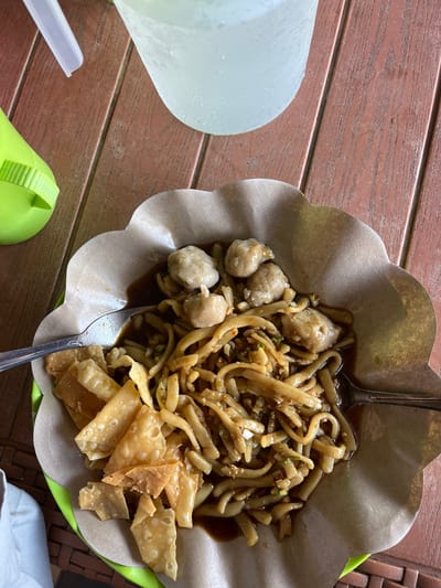 The image displays a close-up, overhead view of a meal served on a scalloped, brown paper liner within a green plastic plate. The meal consists of noodles coated in a dark sauce, several round meatballs, and crispy fried wonton pieces. Two spoons are visible, one partially submerged in the noodles and the other resting on the edge of the plate. In the background, on a wooden table, are a clear plastic cup filled with ice water and a portion of a bright green object, possibly a bottle or dispenser. The wooden table has a textured, planked surface. The context of Welado, Indonesia, is not discernible from the visual information provided in the image.
