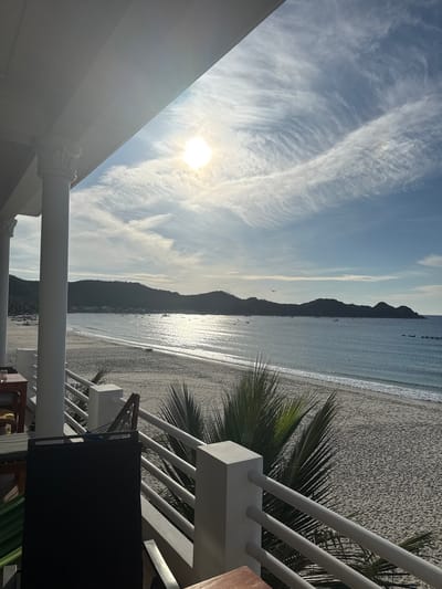 The image depicts a view from a balcony overlooking a beach and the ocean. A white colonnade and railing frame the foreground, with a dark chair and a table partially visible on the left. The beach is sandy and stretches towards a calm, sparkling ocean. In the distance, a dark, tree-covered mountain range forms the horizon. The sky is blue with wispy white clouds, and the sun is high and bright, casting a glare on the water. A few small boats are scattered across the ocean, and a small, dark object, possibly an aircraft or bird, is visible in the sky above the mountains. The scene suggests a serene coastal setting, possibly a resort or a private residence, in Lệ Uyên, Vietnam.