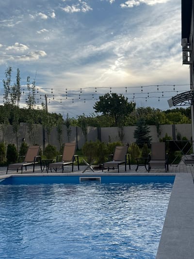 A swimming pool area is depicted, likely in Mughni, Armenia, given the context provided. In the foreground, the blue water of a swimming pool fills the lower half of the frame, with gentle ripples disturbing its surface. Along the pool's edge, a paved area is visible, featuring several lounge chairs. To the left, two brown fabric lounge chairs with dark metal frames are positioned parallel to the pool. Further to the right, two more similar lounge chairs are visible. Between these chairs, small side tables are placed. Behind the lounge chairs, a grey concrete fence runs horizontally across the image, separating the pool area from a backdrop of lush green trees and foliage. String lights are suspended above the fence, adding a decorative element to the scene. A portion of a building with a dark facade is visible on the far right edge of the frame, above the lounge chairs. The sky above is filled with a mix of white clouds and patches of blue.