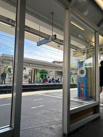The image depicts a daytime scene at the Bourg-la-Reine RER station in France, viewed from within a glass and metal-framed covered area. In the foreground, the concrete ground of a platform features white dashed markings. To the right, a blue and red automated machine, possibly a ticket vending machine or information kiosk, is partially visible and reflects elements of the station. A person in dark attire is partially visible on the far right.
Across a set of train tracks in the midground, an opposing platform is populated by several individuals. On the far left of this platform, a man in a dark jacket and blue jeans stands near the edge. Further right, a group of five individuals sits on a bench under the platform's canopy. One person wears a light pink tracksuit, and a red suitcase rests beside them. A bright green modular shelter or kiosk stands centrally on the opposite platform. Overhead, a black rectangular sign with a white female pictogram and a right-pointing arrow is suspended, indicating facilities. Another sign, partially visible, displays the word "Appel."
The background reveals residential buildings and trees under a clear blue sky with some scattered white clouds. A prominent white building with columns is visible behind the platform structures. The scene is brightly lit, suggesting fair weather.