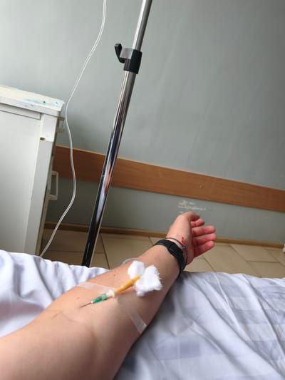In Kovel, Ukraine, an arm lies on a white blanket with an IV drip inserted. The IV tube runs to a pole. A wooden board runs along the wall, above tiled floors. A white cabinet is visible on the left.
