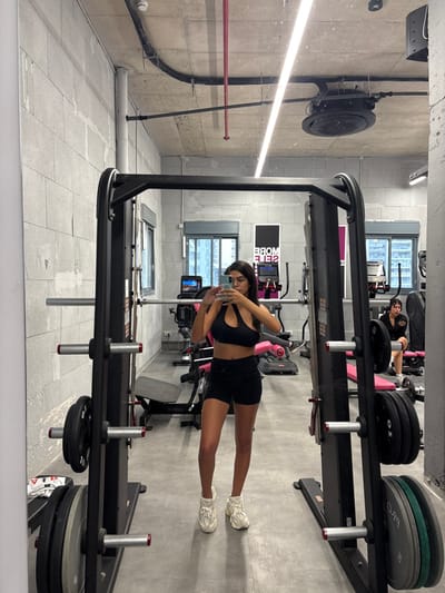 The photo was taken inside a gym located in Ramla, Israel. The gym is well-lit, with a mirrored wall reflecting the scene. A woman in a black sports bra and shorts is standing in front of a weight machine, taking a selfie with her phone. She is wearing white sneakers. The woman is smiling and looking at her phone. There is another woman sitting in the background, also wearing workout attire. The gym is equipped with various weight machines and free weights. The environment is clean and modern. The camera is positioned in a way that the viewer sees the woman taking a selfie through the reflection of the mirrored wall. The photo appears to be taken from a slightly elevated angle. The perspective gives a clear view of the woman’s legs, and the weight machine she is posing in front of.
