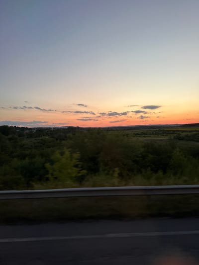 The image depicts a natural landscape at dusk, possibly in or around Pidbirtsi, Ukraine, as indicated. The sky transitions from a pale blue at the top to a warm orange and pink hue near the horizon, illuminated by the setting sun. Scattered clouds are visible against the colorful sky. Below the sky, a treeline with dense green foliage stretches across the mid-ground. Beyond the trees, a field or meadow is visible, with hints of yellow or green crops. In the far distance, the silhouette of buildings and possibly a tall structure, such as a tower or chimney, can be seen, suggesting the presence of a settlement. The foreground features a road with a metallic guardrail, implying the perspective is from a moving vehicle or roadside. The overall scene is peaceful and natural, capturing the tranquil ambiance of twilight. There are no people or animals visible in the image.